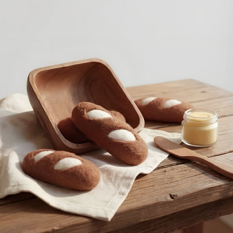 Felt baguettes posed on a wood table accompanied by a wood bowl, spread and a jar of butter